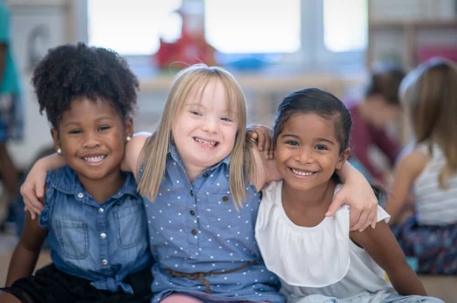 Three little girls sit with arms around one another smiling