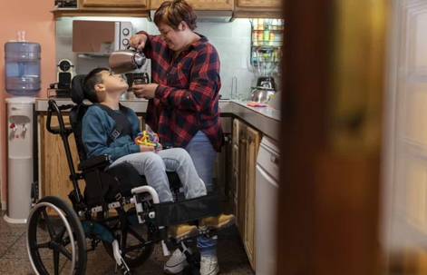 Child with special needs sits in a wheelchair while mom pours tea in the kitchen
