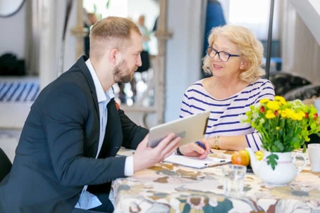 Young businessman sits with a tablet in his hand at a homely table with a flower bouquet with an older woman wearing reading glasses