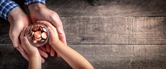 Glass jar filled with pennies sits in the hands of a child with an adult's hands cradling the child's hands over a wooden table