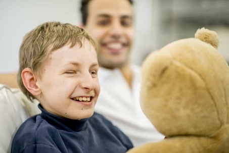 Young boy with special needs holds and smiles at a teddy bear with his father behind him