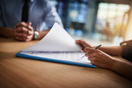 Close up of a woman's hands about to sign documents attached to a clipboard with a businessman sitting at the table across from her