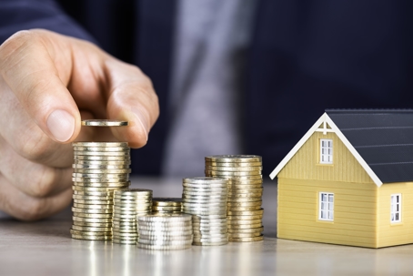 person stacking up gold coins next to a small model of a house